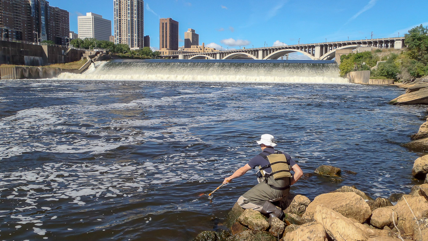 An MWMO staff member wearing safety gear collects a water sample from below St. Anthony Falls.